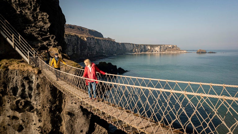 Two visitors walking along Carrick-a-Rede Rope Bridge. Visitor at the front looking down to the Atlantic Ocean. Visitor at the back looking ahead to the island.